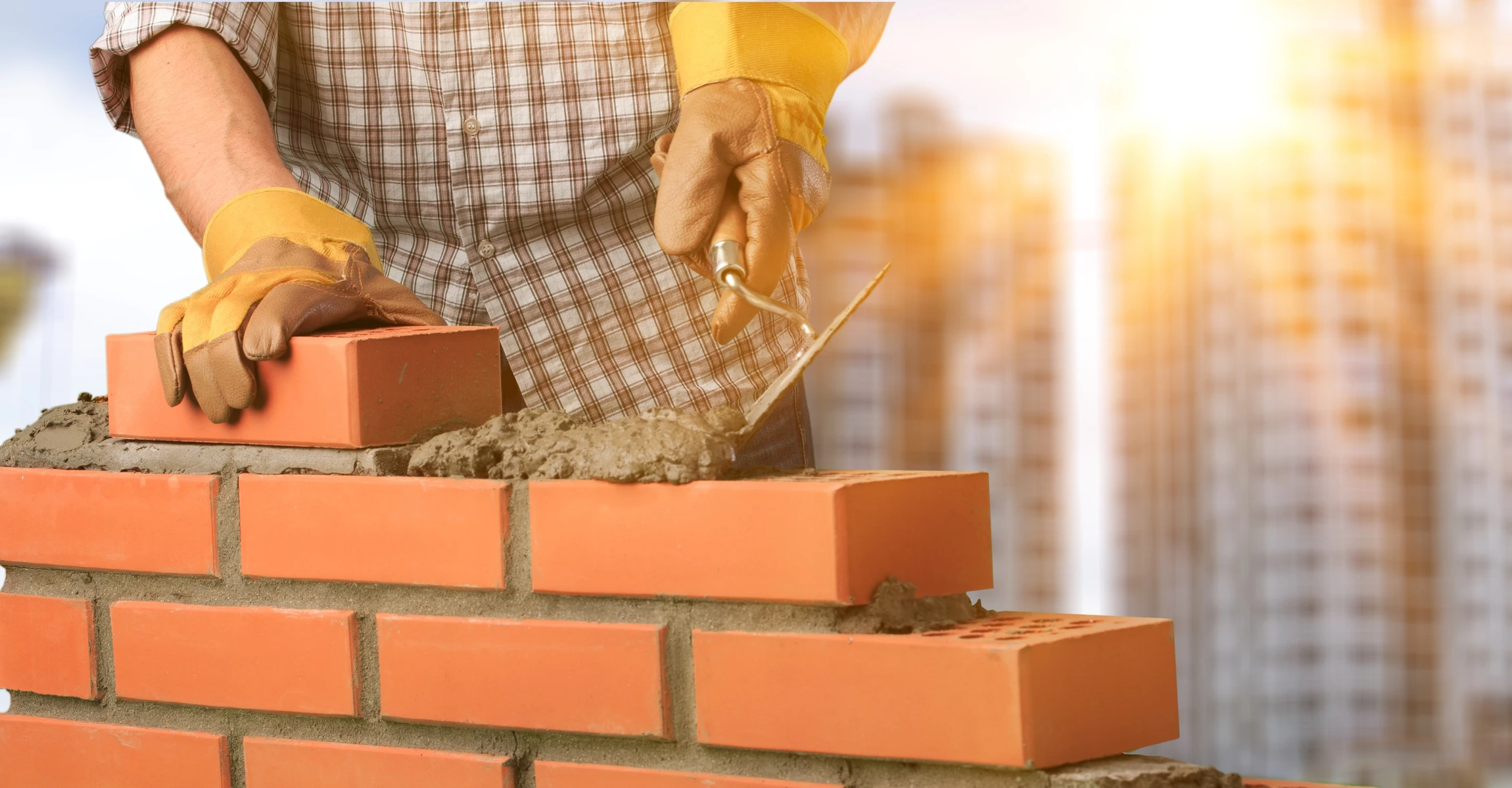 Ouvrier maçon construisant un mur en briques rouges sur chantier extérieur, portant gants de protection jaunes et chemise à carreaux